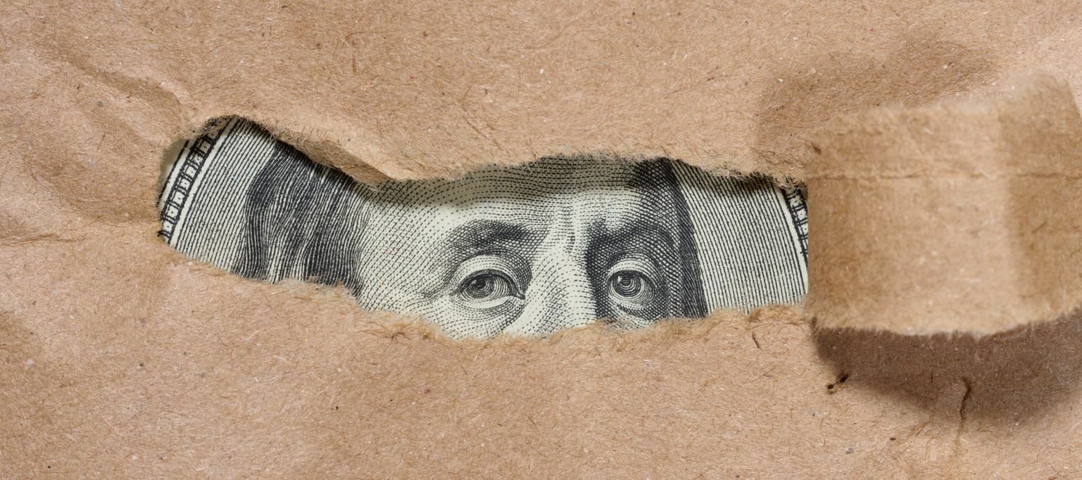 A piece of a hundred dollar bill is visible through a hole in the paper. Symbolism of hidden or concealed wealth, money that remains out of sight, or financial resources.